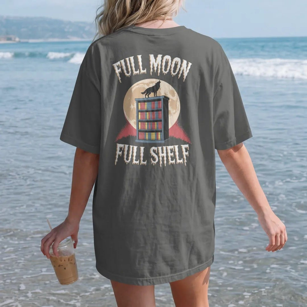 Woman on the beach in a Pepper tee, back view: a full moon behind a bookshelf on a hill with a wolf howling on top, with Full Moon above and Full Shelf below, in pulp halftone cream, red, yellow and blue.
