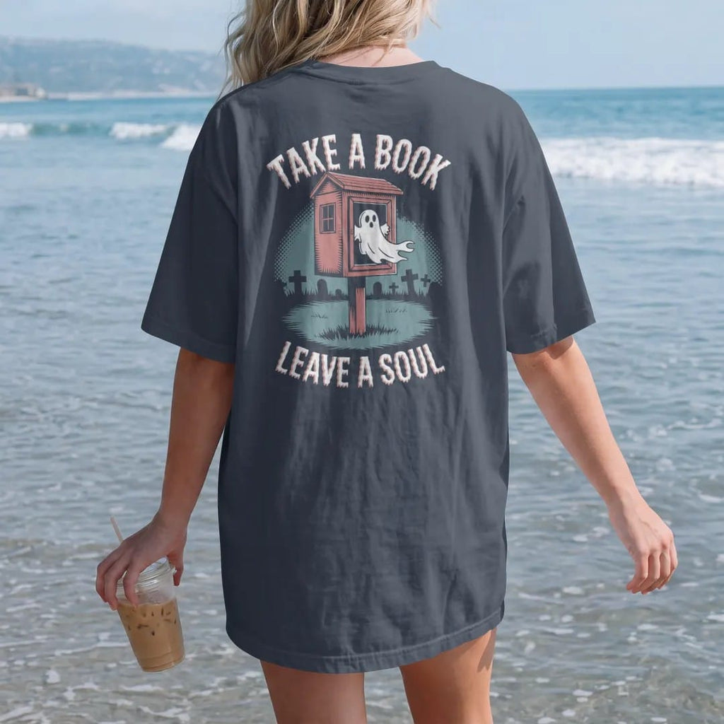Woman on the beach in a Midnight tee, back view: a little free library box in a misty cemetery with a ghost drifting out, Take A Book above and Leave A Soul below, in pulp halftone navy, red, mustard and teal.