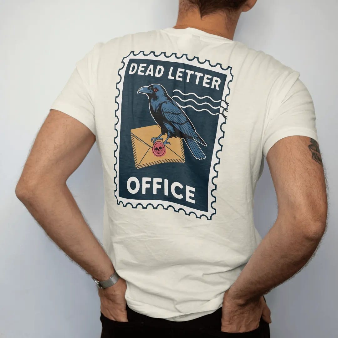 Man wearing a Ivory tee, seen from the back: a vintage postage stamp of a raven carrying a skull-sealed envelope, Dead Letter on top and Office below, in pulp halftone navy, red, mustard and teal.