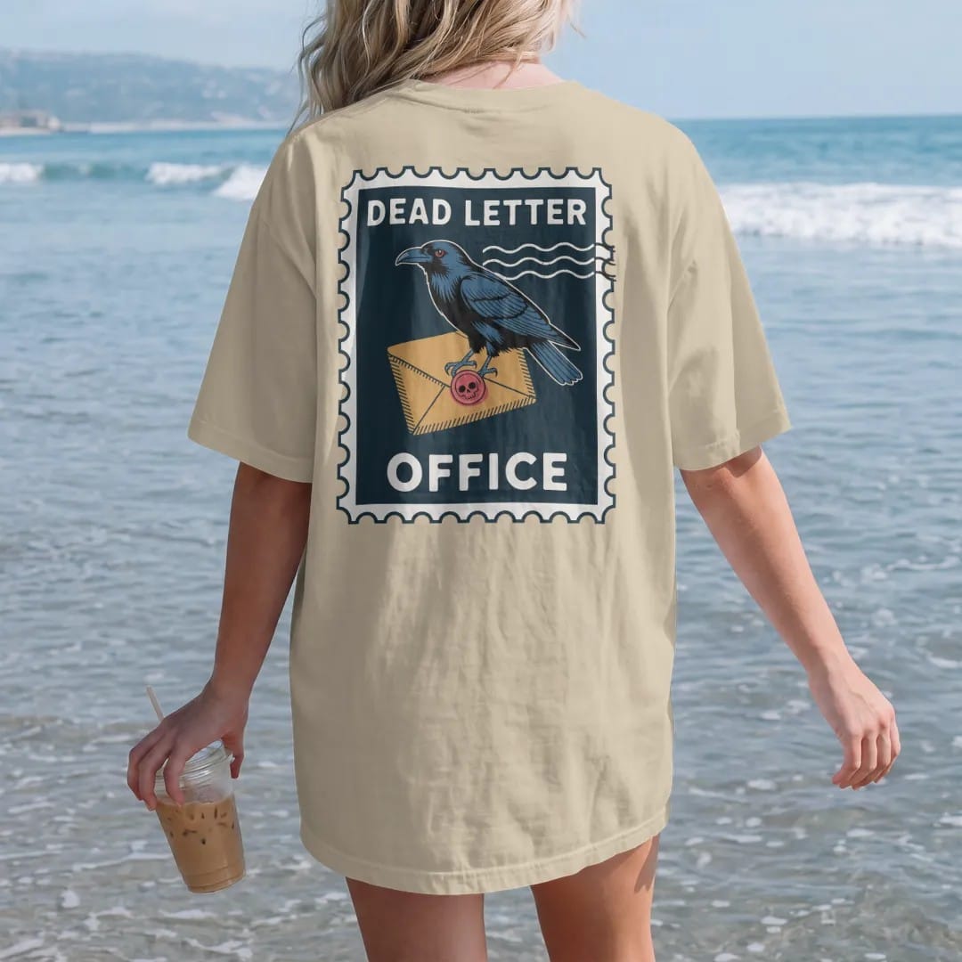 Woman on the beach in a Sandstone tee, back view: a vintage postage stamp of a raven carrying a skull-sealed envelope, Dead Letter on top and Office below, in pulp halftone navy, red, mustard and teal.