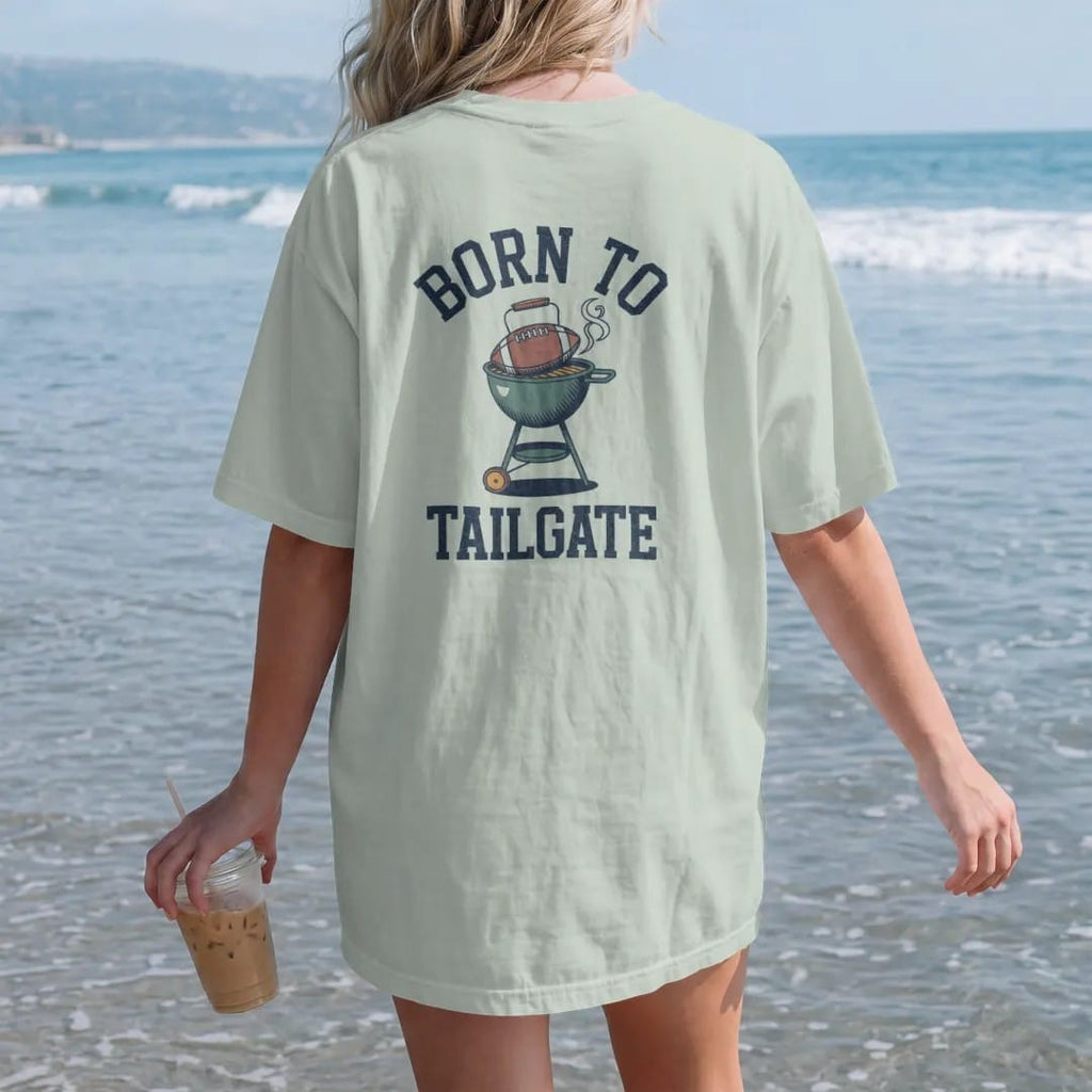 Woman on the beach in a Bay tee, back view: a kettle grill with a football on top, wearing sunglasses, and smoke curling up, with "BORN TO" and "TAILGATE" in chunky 1970s swash script, printed in faded stadium green, washed brick red, faded navy…