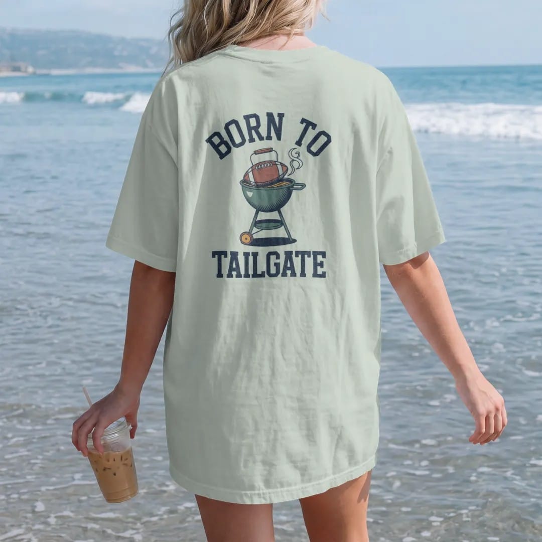 Woman on the beach in a Bay tee, back view: a kettle grill with a football on top, wearing sunglasses, and smoke curling up, with "BORN TO" and "TAILGATE" in chunky 1970s swash script, printed in faded stadium green, washed brick red, faded navy…