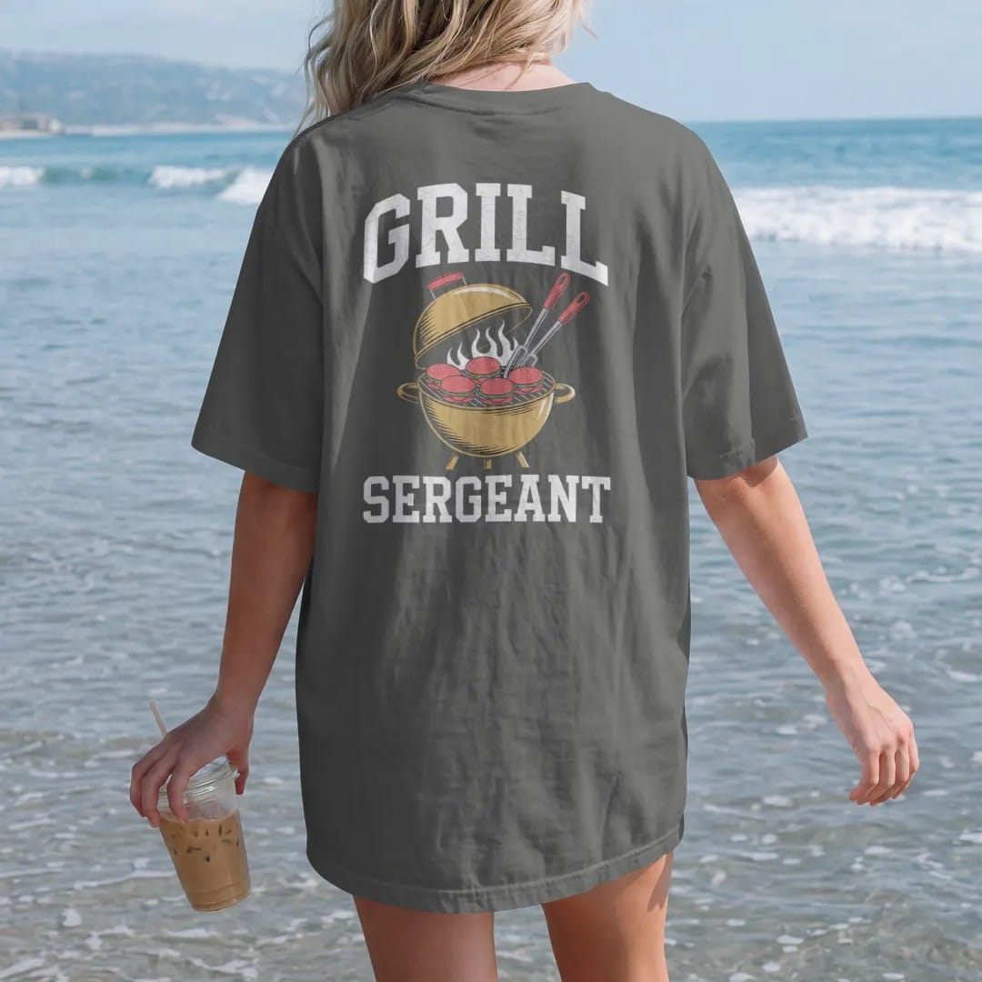 Woman on the beach in a Pepper tee, back view: a spatula and tongs crossed over a flaming kettle grill full of burgers, with "GRILL" and "SERGEANT" in chunky rounded 70s varsity letters, printed in faded bleacher cream, mustard, washed tomato red…