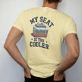 Man wearing a Butter tee, seen from the back: a classic two-tone cooler with a stadium cushion on top and cans on ice, with "MY SEAT" and "IS THE COOLER" in chunky 70s slab-serif letters, printed in faded stadium green, washed brick red, faded navy…