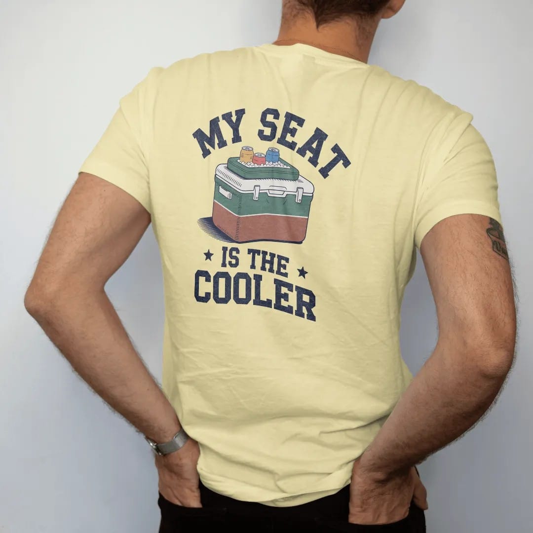 Man wearing a Butter tee, seen from the back: a classic two-tone cooler with a stadium cushion on top and cans on ice, with "MY SEAT" and "IS THE COOLER" in chunky 70s slab-serif letters, printed in faded stadium green, washed brick red, faded navy…
