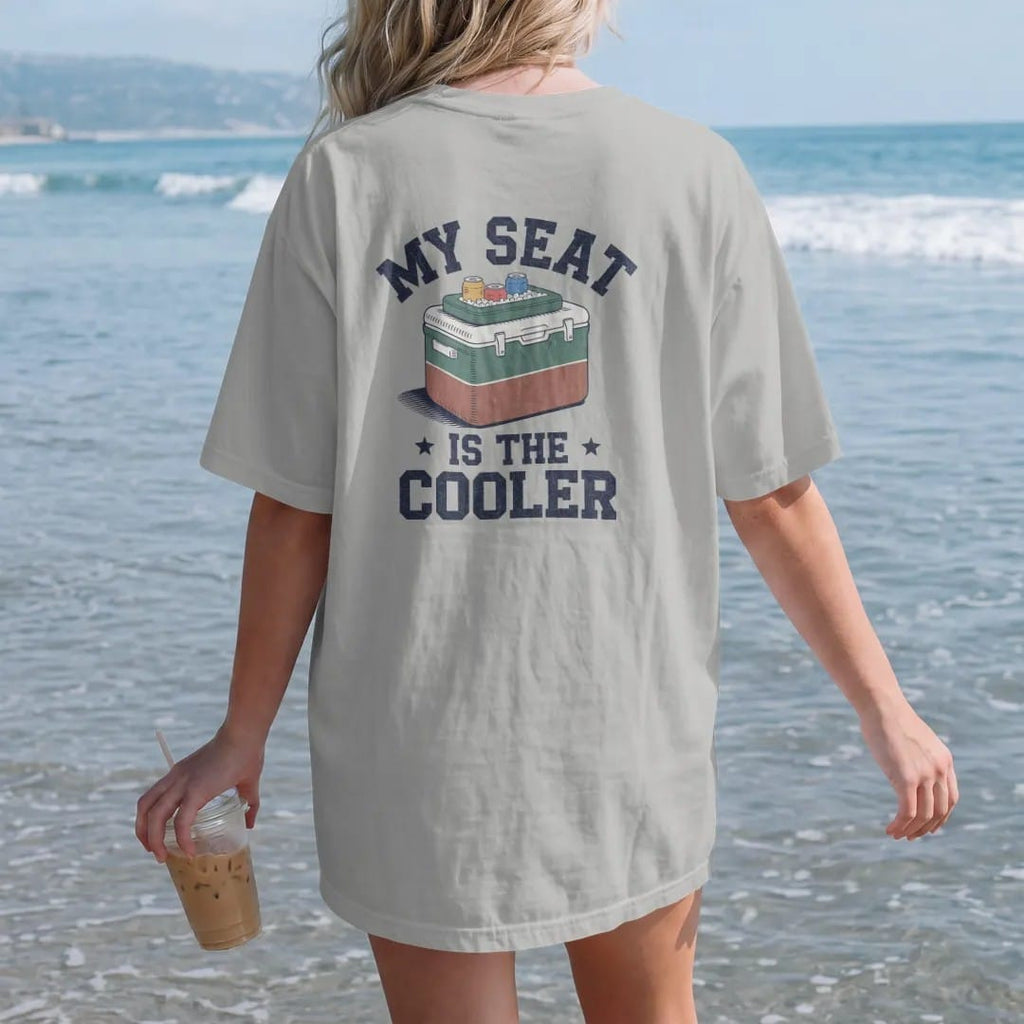 Woman on the beach in a Granite tee, back view: a classic two-tone cooler with a stadium cushion on top and cans on ice, with "MY SEAT" and "IS THE COOLER" in chunky 70s slab-serif letters, printed in faded stadium green, washed brick red, faded…