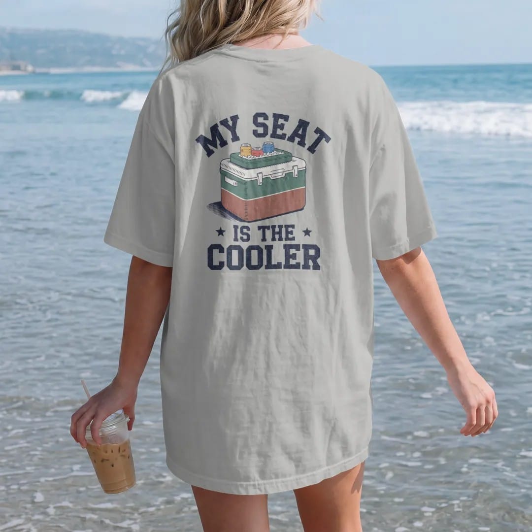 Woman on the beach in a Granite tee, back view: a classic two-tone cooler with a stadium cushion on top and cans on ice, with "MY SEAT" and "IS THE COOLER" in chunky 70s slab-serif letters, printed in faded stadium green, washed brick red, faded…