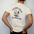 Man wearing a Ivory tee, seen from the back: a striped folding camp chair with a cup holder and a foam finger, with "FOLDING CHAIR" and "ALL-AMERICAN" in chunky rounded 70s varsity letters, printed in faded stadium green, washed brick red, faded…