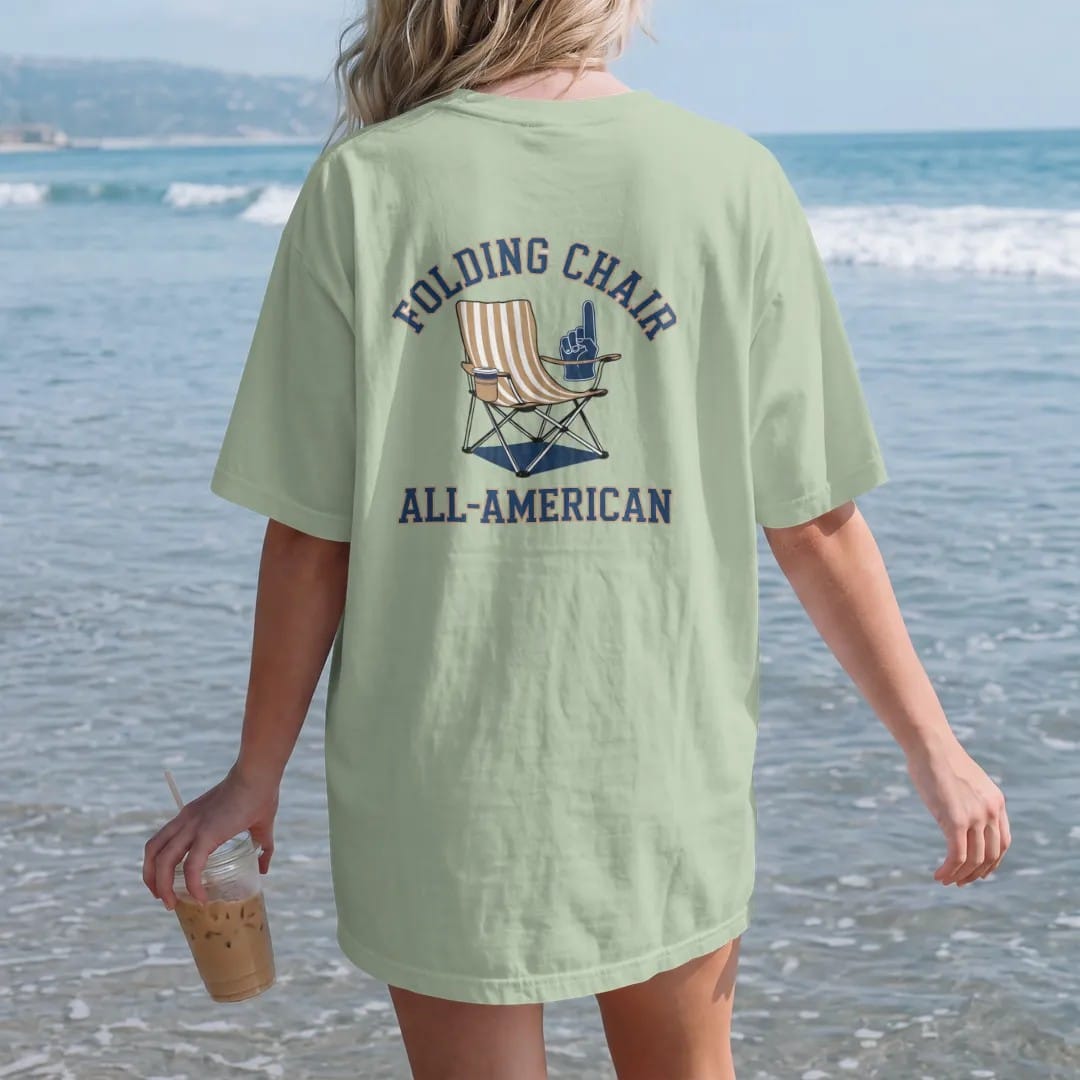 Woman on the beach in a Light Green tee, back view: a striped folding camp chair with a cup holder and a foam finger, with "FOLDING CHAIR" and "ALL-AMERICAN" in chunky rounded 70s varsity letters, printed in faded stadium green, washed brick red,…