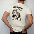 Man wearing a Ivory tee, seen from the back: a vintage alarm clock with a football face beside a steaming coffee mug, with "KICKOFF AT NOON" and "TAILGATE AT 7" in chunky 70s slab-serif letters, printed in faded stadium green, washed brick red,…
