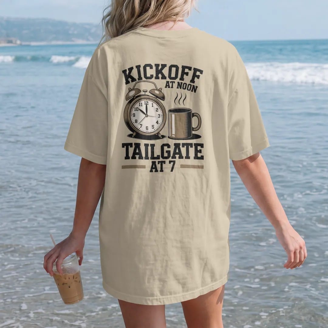 Woman on the beach in a Sandstone tee, back view: a vintage alarm clock with a football face beside a steaming coffee mug, with "KICKOFF AT NOON" and "TAILGATE AT 7" in chunky 70s slab-serif letters, printed in faded stadium green, washed brick…