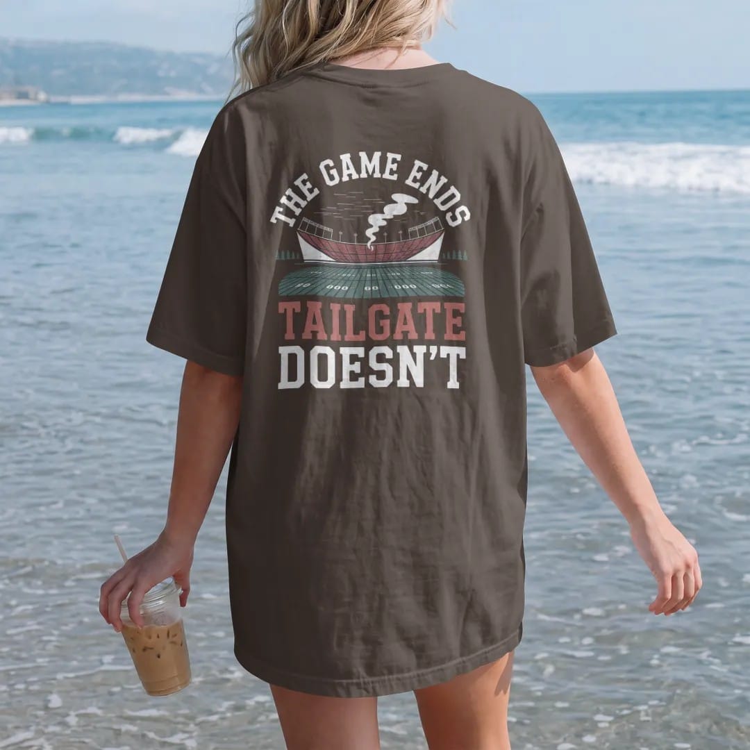 Woman on the beach in a Espresso tee, back view: a stadium at dusk with the lights on and grill smoke drifting over the parking lot, with "THE GAME ENDS" and "THE TAILGATE DOESN'T" in chunky rounded 70s varsity letters, printed in faded bleacher…