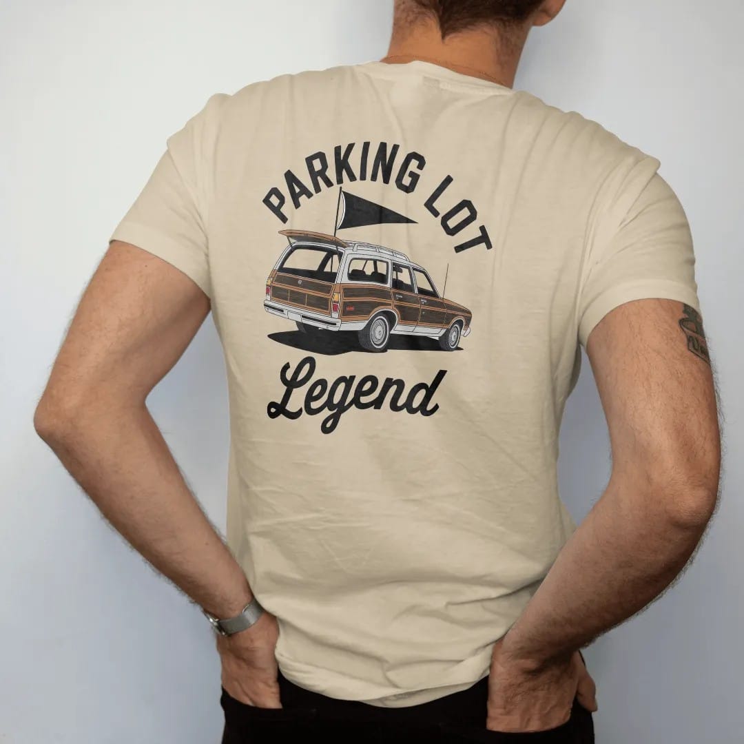 Man wearing a Sandstone tee, seen from the back: a classic wood-paneled station wagon with the tailgate down, a grill and a pennant flag, with "PARKING LOT" and "LEGEND" in chunky 1970s swash script, printed in faded stadium green, washed brick…