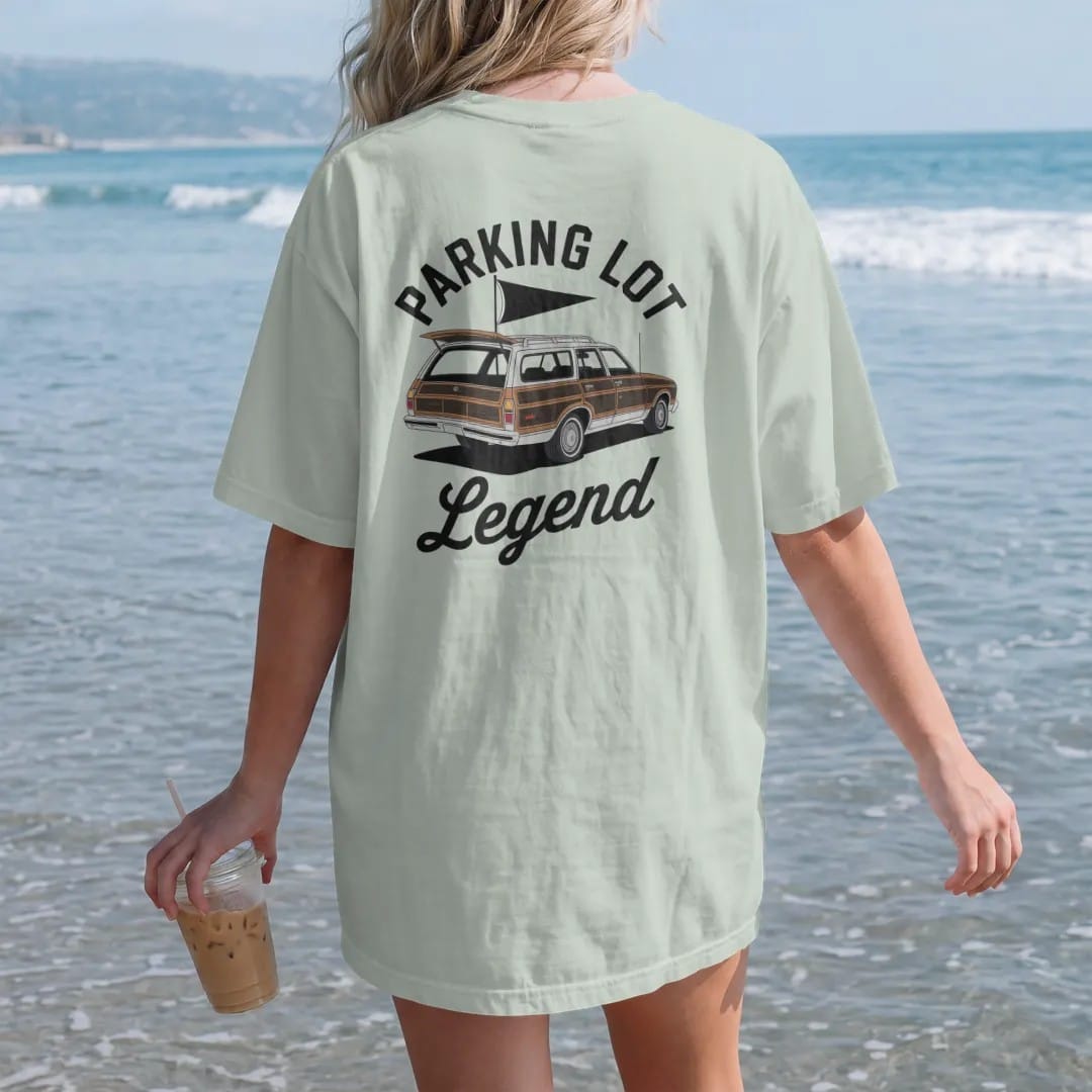 Woman on the beach in a Bay tee, back view: a classic wood-paneled station wagon with the tailgate down, a grill and a pennant flag, with "PARKING LOT" and "LEGEND" in chunky 1970s swash script, printed in faded stadium green, washed brick red,…