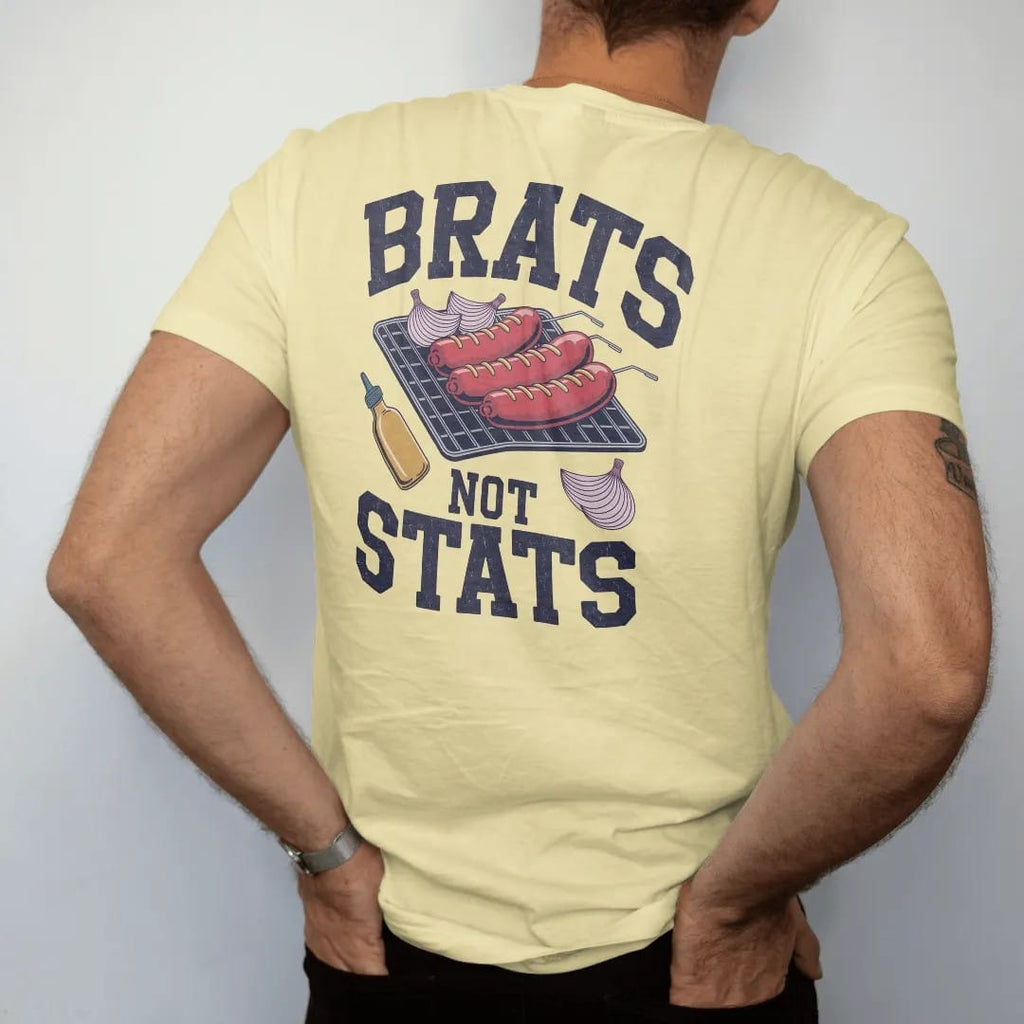 Man wearing a Butter tee, seen from the back: sizzling bratwursts on grill grates with onions and a mustard bottle, with "BRATS" and "NOT STATS" in chunky rounded 70s varsity letters, printed in faded stadium green, washed brick red, faded navy and…