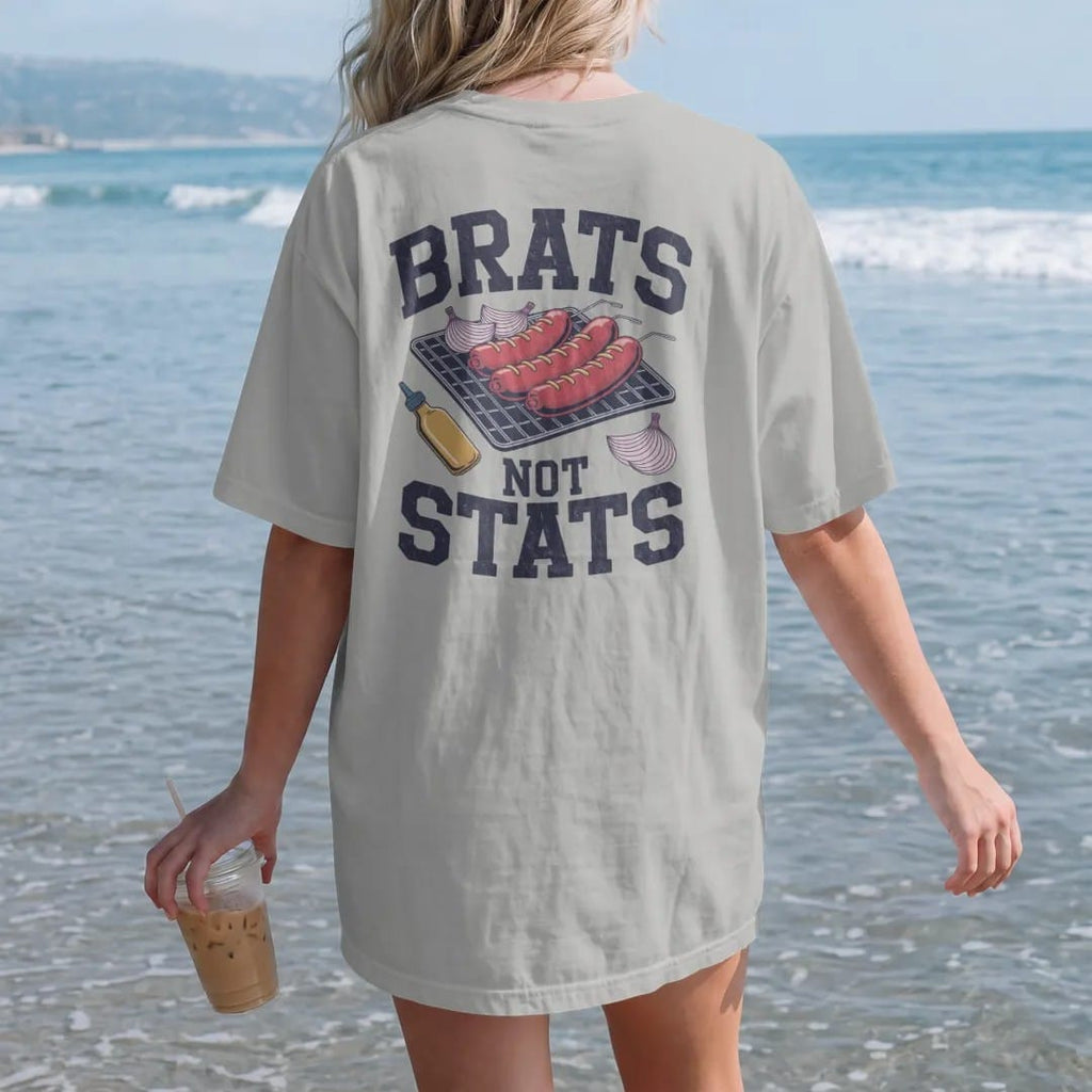 Woman on the beach in a Granite tee, back view: sizzling bratwursts on grill grates with onions and a mustard bottle, with "BRATS" and "NOT STATS" in chunky rounded 70s varsity letters, printed in faded stadium green, washed brick red, faded navy…