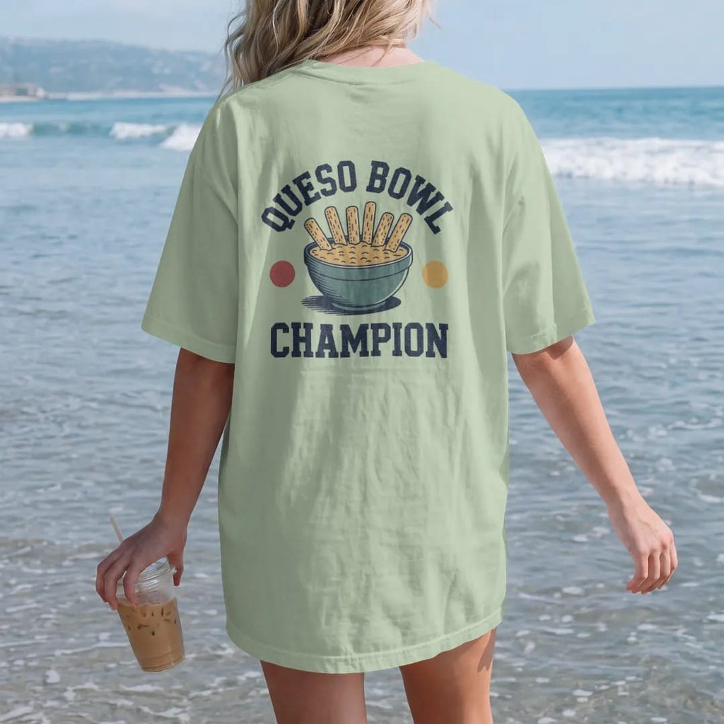 Woman on the beach in a Light Green tee, back view: a giant bowl of queso with tortilla chips standing up like goalposts, with "QUESO BOWL" and "CHAMPION" in chunky 70s slab-serif letters, printed in faded stadium green, washed brick red, faded…