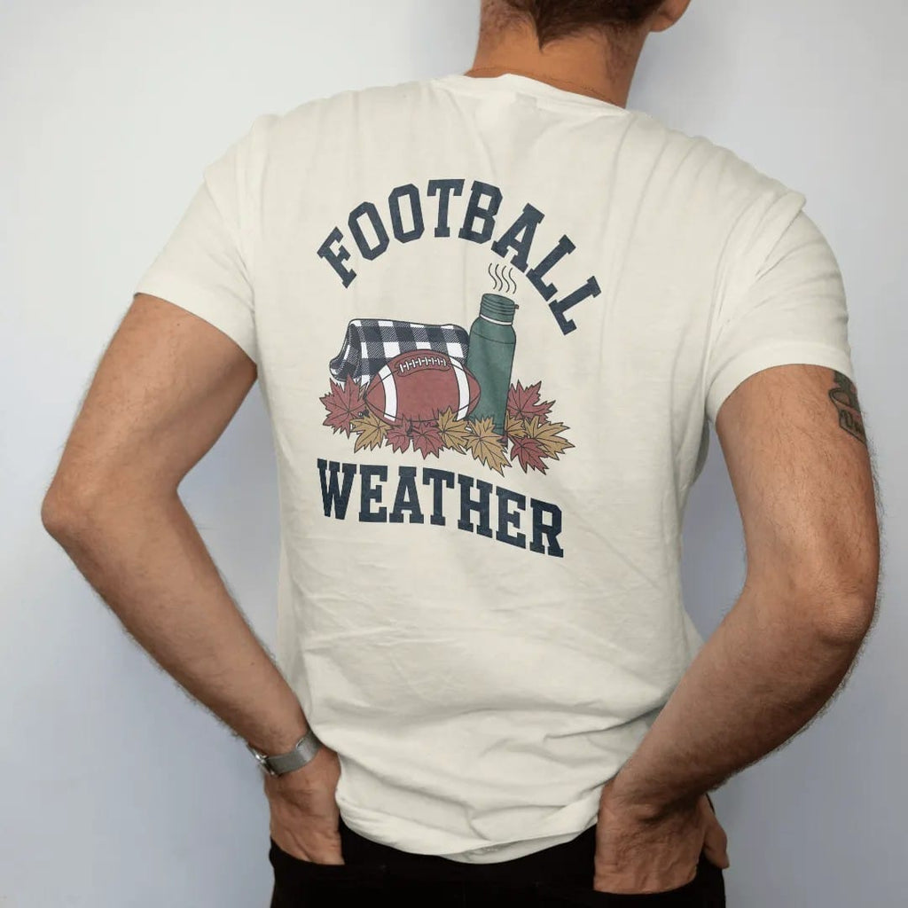 Man wearing a Ivory tee, seen from the back: a football in a pile of autumn leaves with a steaming thermos and a flannel blanket, with "FOOTBALL WEATHER" in chunky 1970s swash script, printed in faded stadium green, washed brick red, faded navy and…