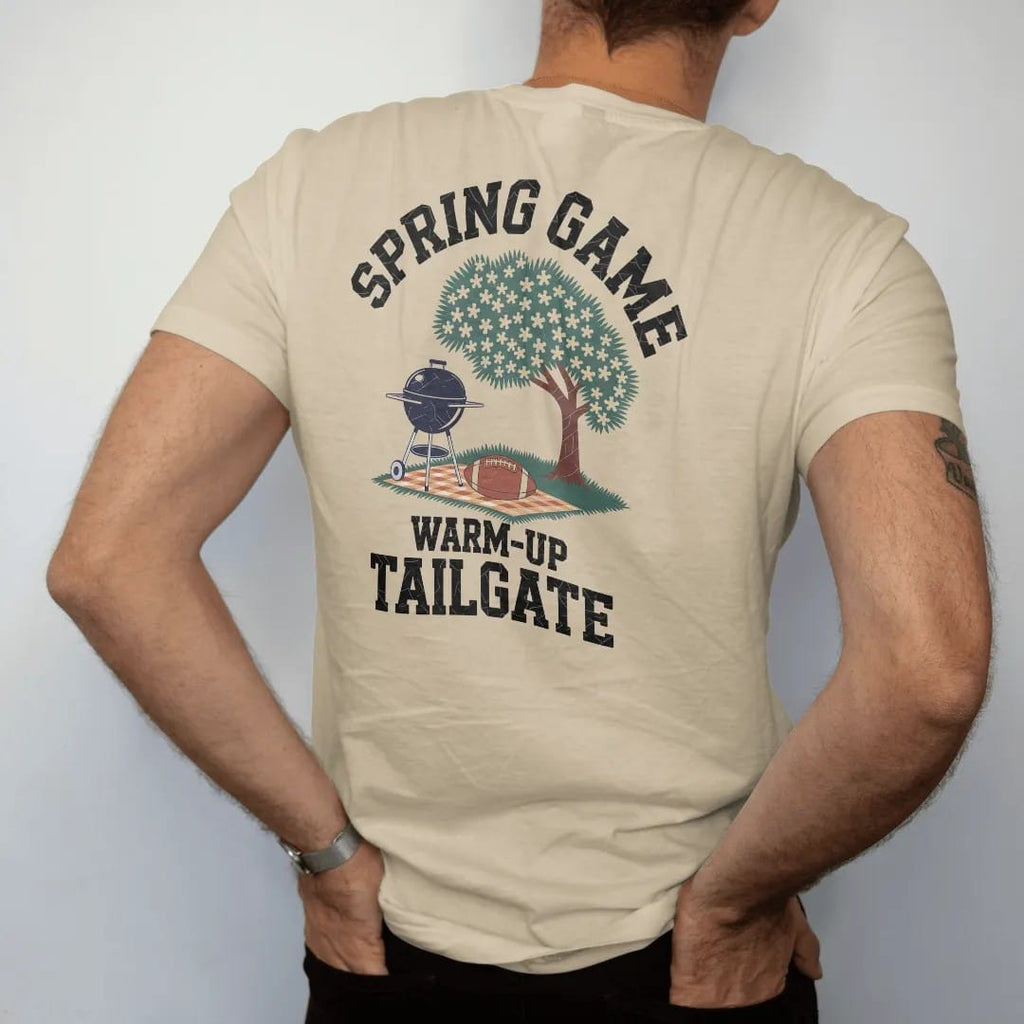 Man wearing a Sandstone tee, seen from the back: a grill under a blooming dogwood tree with a football on a picnic blanket, with "SPRING GAME" and "WARM-UP TAILGATE" in chunky rounded 70s varsity letters, printed in faded stadium green, washed…