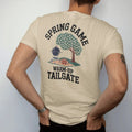 Man wearing a Sandstone tee, seen from the back: a grill under a blooming dogwood tree with a football on a picnic blanket, with "SPRING GAME" and "WARM-UP TAILGATE" in chunky rounded 70s varsity letters, printed in faded stadium green, washed…