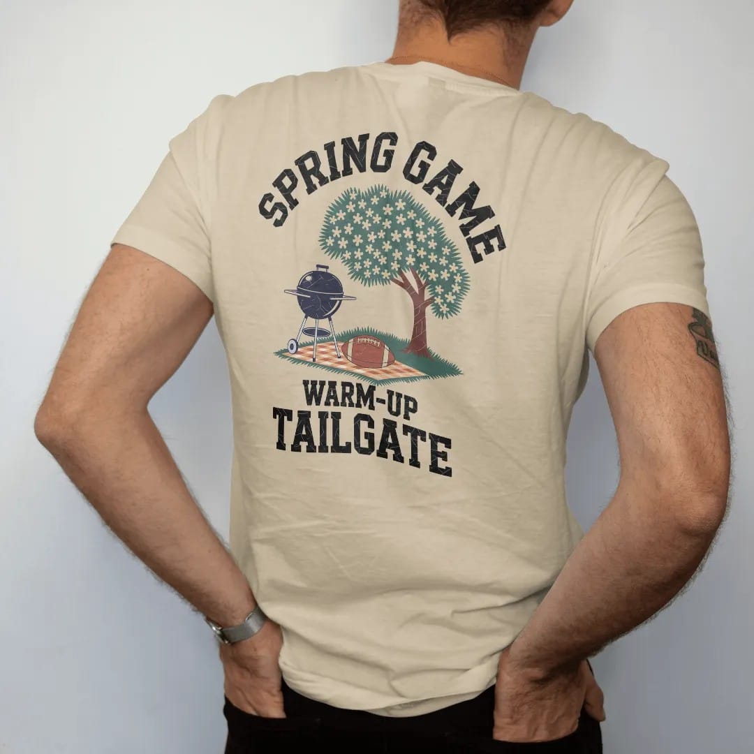 Man wearing a Sandstone tee, seen from the back: a grill under a blooming dogwood tree with a football on a picnic blanket, with "SPRING GAME" and "WARM-UP TAILGATE" in chunky rounded 70s varsity letters, printed in faded stadium green, washed…