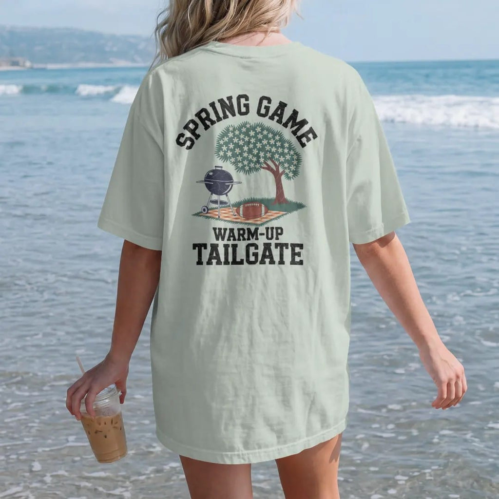 Woman on the beach in a Bay tee, back view: a grill under a blooming dogwood tree with a football on a picnic blanket, with "SPRING GAME" and "WARM-UP TAILGATE" in chunky rounded 70s varsity letters, printed in faded stadium green, washed brick…