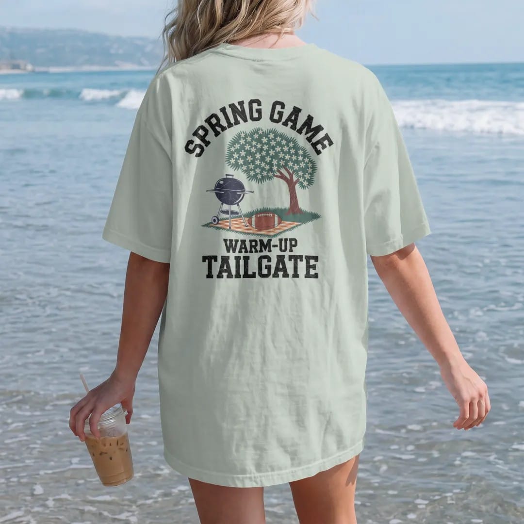 Woman on the beach in a Bay tee, back view: a grill under a blooming dogwood tree with a football on a picnic blanket, with "SPRING GAME" and "WARM-UP TAILGATE" in chunky rounded 70s varsity letters, printed in faded stadium green, washed brick…