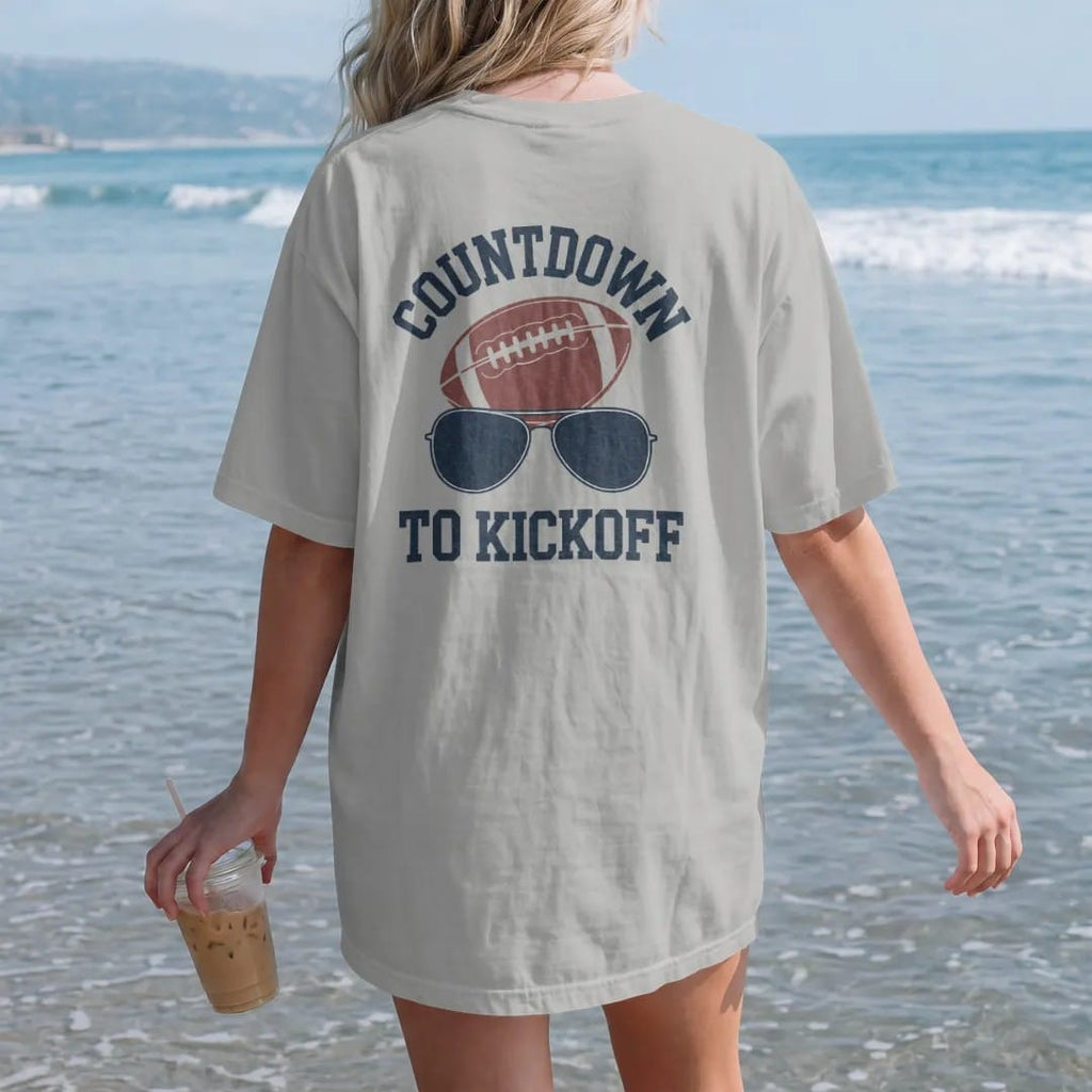 Woman on the beach in a Granite tee, back view: a sun-faded calendar page with days crossed off beside a football and sunglasses, with "COUNTDOWN" and "TO KICKOFF" in chunky 70s slab-serif letters, printed in faded stadium green, washed brick red,…