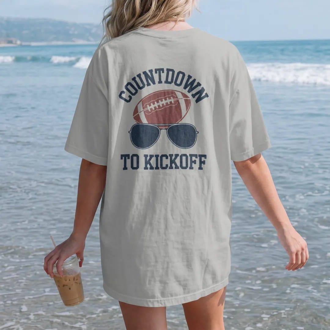 Woman on the beach in a Granite tee, back view: a sun-faded calendar page with days crossed off beside a football and sunglasses, with "COUNTDOWN" and "TO KICKOFF" in chunky 70s slab-serif letters, printed in faded stadium green, washed brick red,…