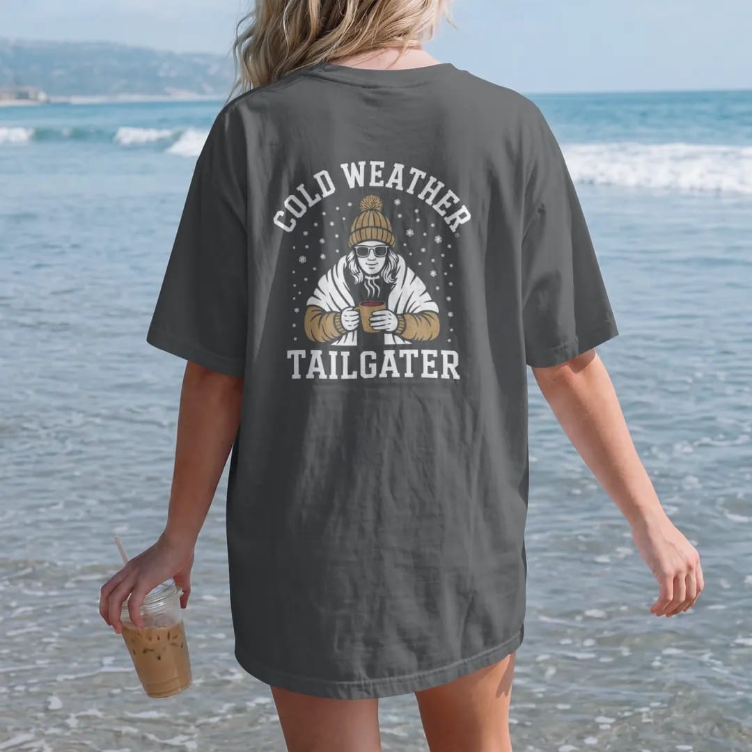 Woman on the beach in a Graphite tee, back view: a fan bundled in a knit pom hat and blanket holding a steaming cup of chili as snow falls, with "COLD WEATHER" and "TAILGATER" in chunky rounded 70s varsity letters, printed in faded bleacher cream,…