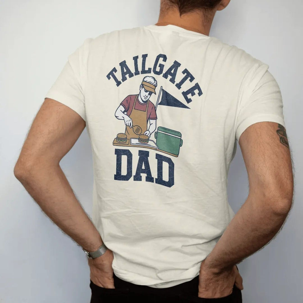Man wearing a Ivory tee, seen from the back: a dad in a visor flipping burgers, a cooler and a pennant behind him, with "TAILGATE" and "DAD" in chunky 1970s swash script, printed in faded stadium green, washed brick red, faded navy and mustard with…