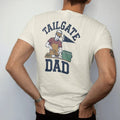 Man wearing a Ivory tee, seen from the back: a dad in a visor flipping burgers, a cooler and a pennant behind him, with "TAILGATE" and "DAD" in chunky 1970s swash script, printed in faded stadium green, washed brick red, faded navy and mustard with…