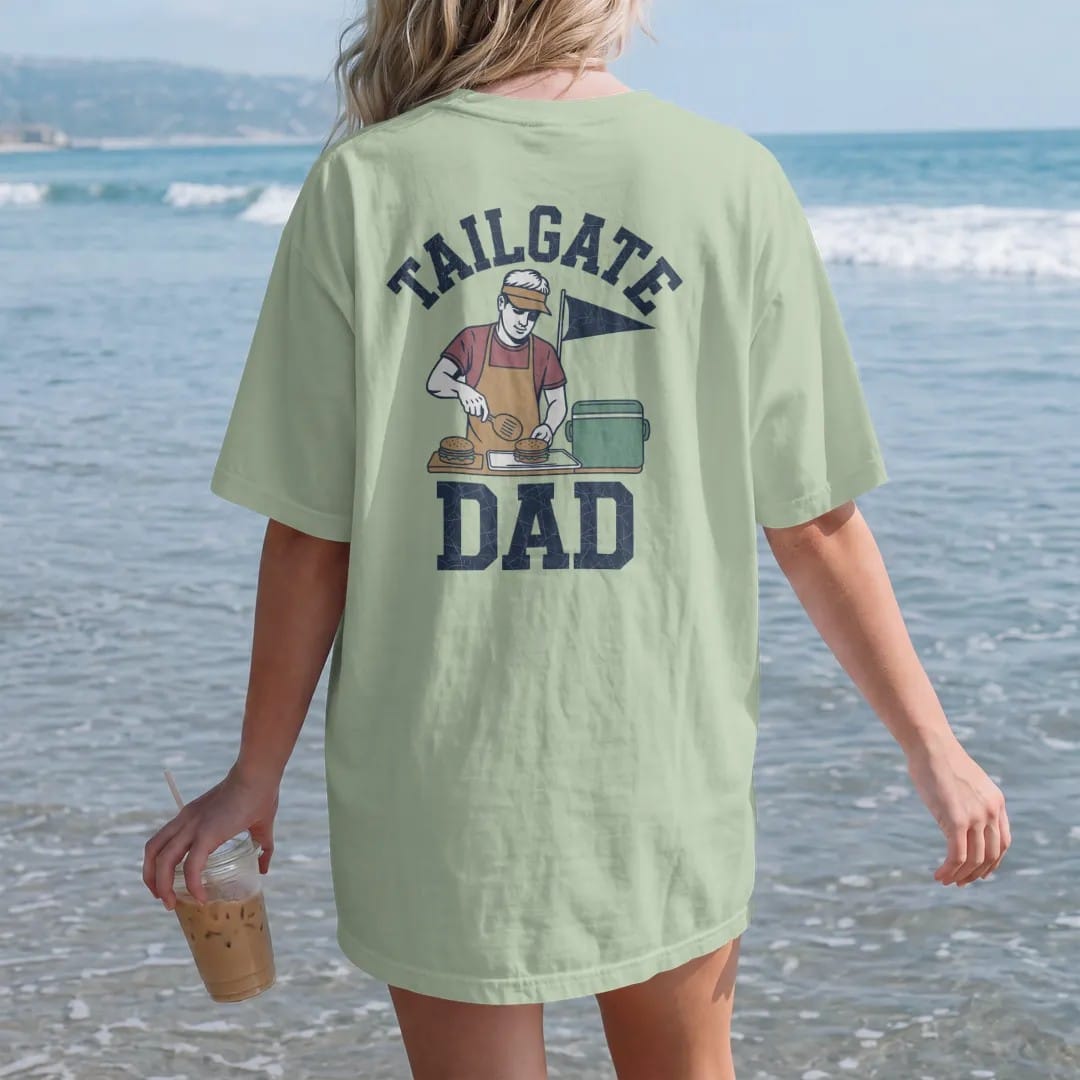 Woman on the beach in a Light Green tee, back view: a dad in a visor flipping burgers, a cooler and a pennant behind him, with "TAILGATE" and "DAD" in chunky 1970s swash script, printed in faded stadium green, washed brick red, faded navy and…