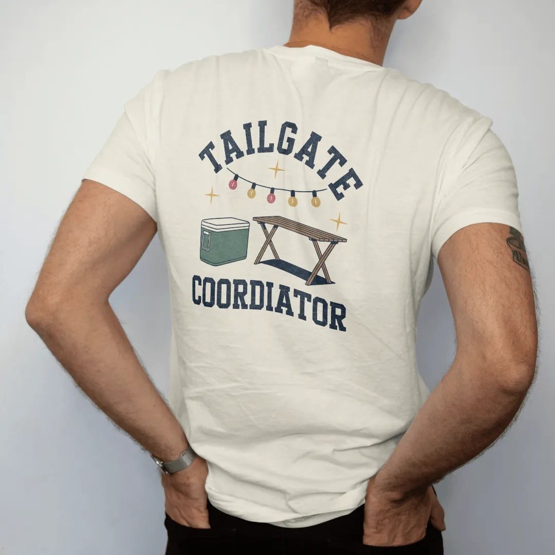 Man wearing a Ivory tee, seen from the back: a clipboard checklist beside a cooler, a folding table and string lights, with "TAILGATE" and "COORDINATOR" in chunky 70s slab-serif letters, printed in faded stadium green, washed brick red, faded navy…