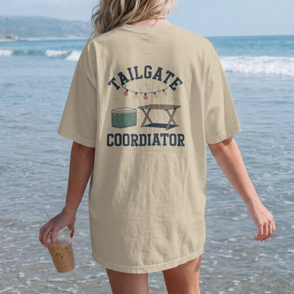 Woman on the beach in a Sandstone tee, back view: a clipboard checklist beside a cooler, a folding table and string lights, with "TAILGATE" and "COORDINATOR" in chunky 70s slab-serif letters, printed in faded stadium green, washed brick red, faded…