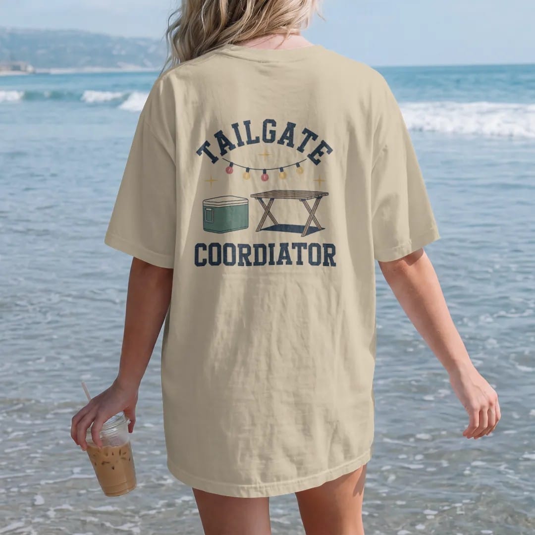 Woman on the beach in a Sandstone tee, back view: a clipboard checklist beside a cooler, a folding table and string lights, with "TAILGATE" and "COORDINATOR" in chunky 70s slab-serif letters, printed in faded stadium green, washed brick red, faded…