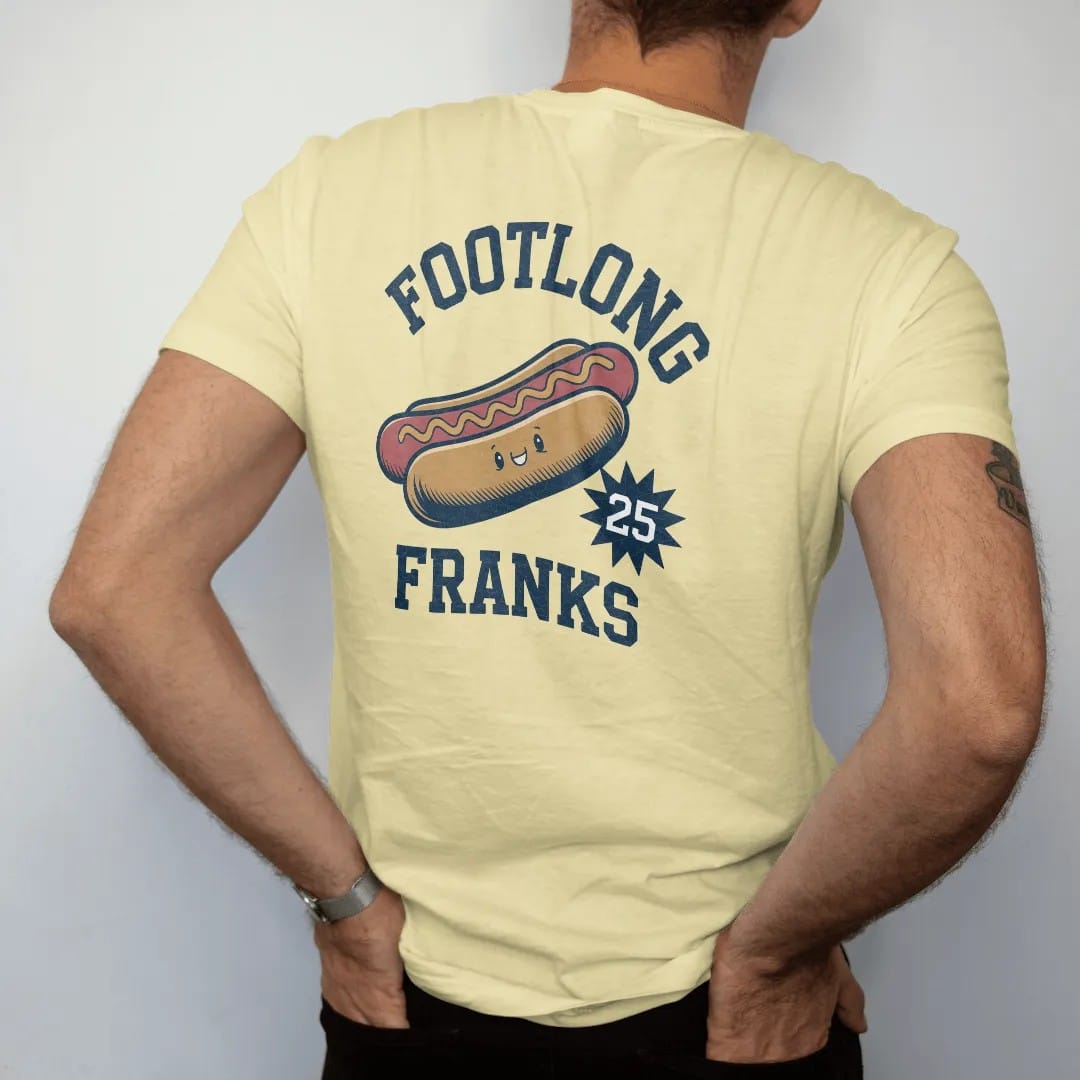 Man wearing a Butter tee, seen from the back: a vintage concession hot dog with a mustard zigzag and a cartoon smiling face, with "FOOTLONG FRANKS" and "25 CENTS" in chunky rounded 70s sign letters, printed in faded stadium green, washed brick red,…