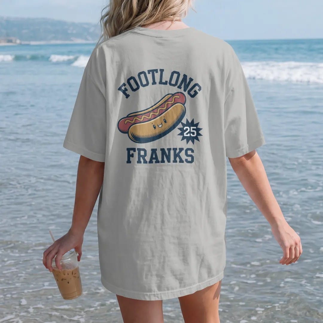 Woman on the beach in a Granite tee, back view: a vintage concession hot dog with a mustard zigzag and a cartoon smiling face, with "FOOTLONG FRANKS" and "25 CENTS" in chunky rounded 70s sign letters, printed in faded stadium green, washed brick…