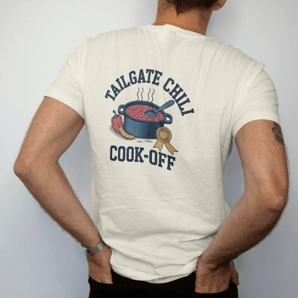 Man wearing a Ivory tee, seen from the back: a cast-iron pot of bubbling chili with a ladle, peppers and a prize ribbon, with "TAILGATE CHILI" and "COOK-OFF" in chunky rounded 70s varsity letters, printed in faded stadium green, washed brick red,…