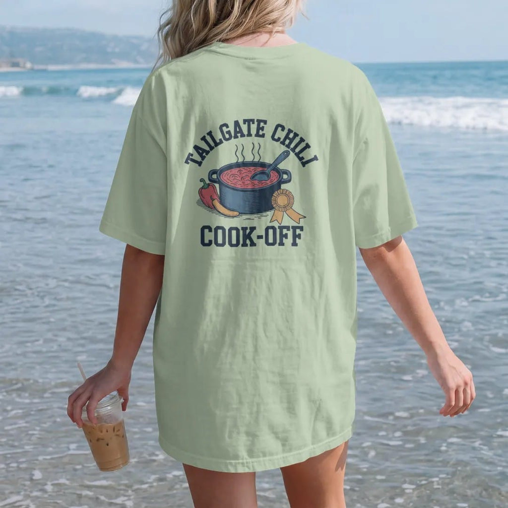 Woman on the beach in a Light Green tee, back view: a cast-iron pot of bubbling chili with a ladle, peppers and a prize ribbon, with "TAILGATE CHILI" and "COOK-OFF" in chunky rounded 70s varsity letters, printed in faded stadium green, washed brick…