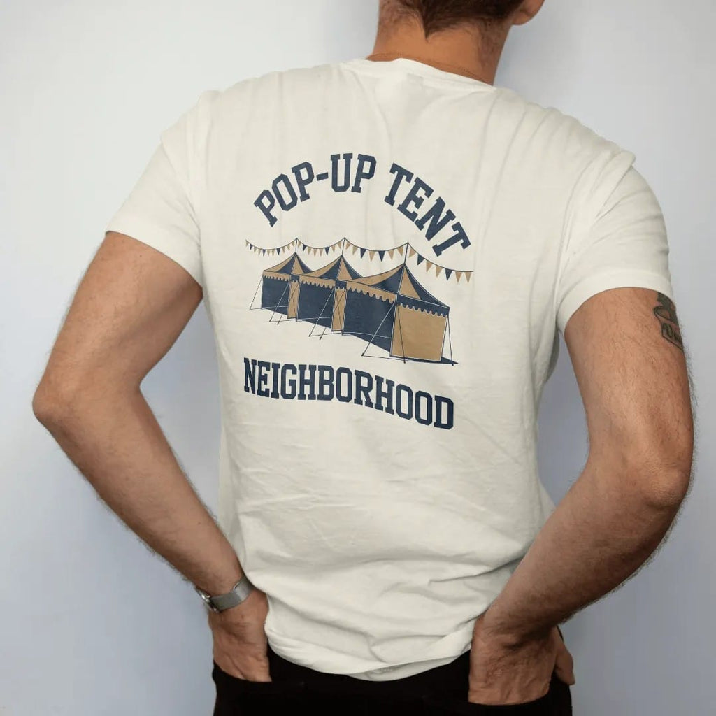 Man wearing a Ivory tee, seen from the back: a row of pop-up canopy tents with pennant strings and flags, with "POP-UP TENT" and "NEIGHBORHOOD" in chunky 70s slab-serif letters, printed in faded stadium green, washed brick red, faded navy and…