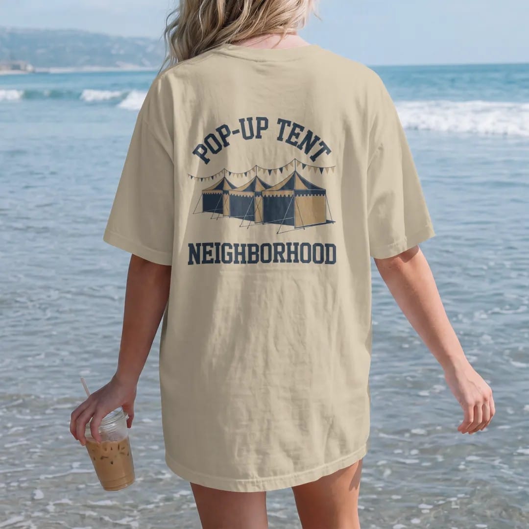 Woman on the beach in a Sandstone tee, back view: a row of pop-up canopy tents with pennant strings and flags, with "POP-UP TENT" and "NEIGHBORHOOD" in chunky 70s slab-serif letters, printed in faded stadium green, washed brick red, faded navy and…