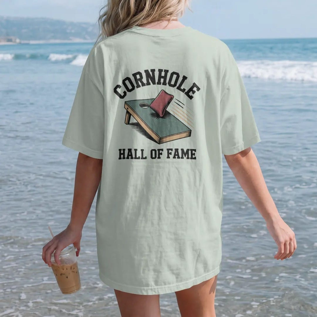 Woman on the beach in a Bay tee, back view: a cornhole board with a bean bag dropping into the hole with motion lines, with "CORNHOLE" and "HALL OF FAME" in chunky rounded 70s varsity letters, printed in faded stadium green, washed brick red, faded…