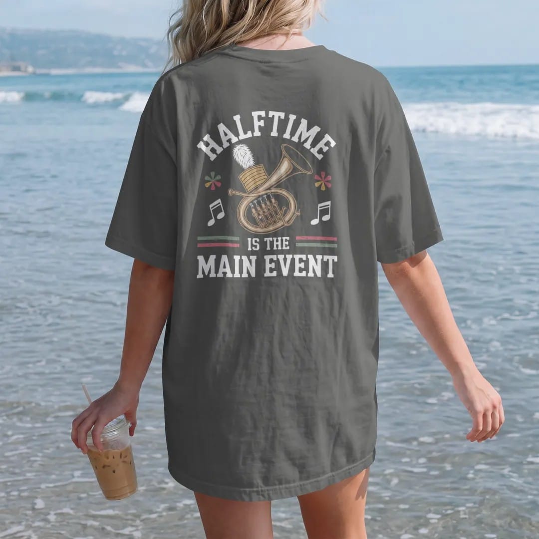 Woman on the beach in a Pepper tee, back view: a marching band sousaphone with a plumed hat and musical notes, with "HALFTIME" and "IS THE MAIN EVENT" in chunky 1970s swash script, printed in faded bleacher cream, mustard, washed tomato red and…