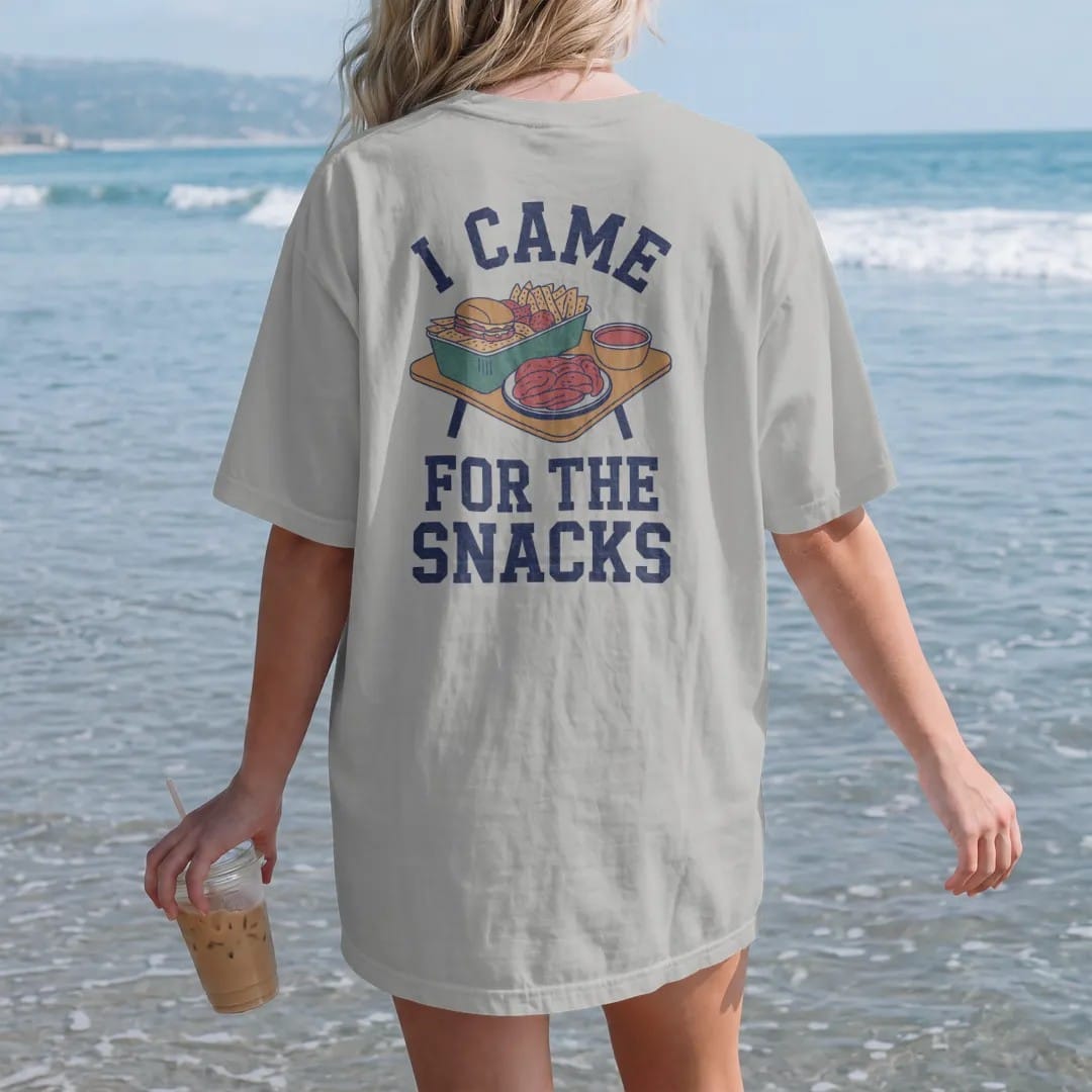 Woman on the beach in a Granite tee, back view: a loaded tailgate table with sliders, chips, dips and wings, with "I CAME" and "FOR THE SNACKS" in chunky 70s slab-serif letters, printed in faded stadium green, washed brick red, faded navy and…