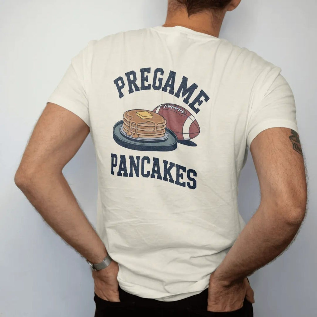 Man wearing a Ivory tee, seen from the back: a stack of pancakes on a camp griddle with butter and syrup, a football beside them, with "PREGAME" and "PANCAKES" in chunky 1970s swash script, printed in faded stadium green, washed brick red, faded…