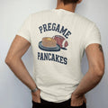 Man wearing a Ivory tee, seen from the back: a stack of pancakes on a camp griddle with butter and syrup, a football beside them, with "PREGAME" and "PANCAKES" in chunky 1970s swash script, printed in faded stadium green, washed brick red, faded…