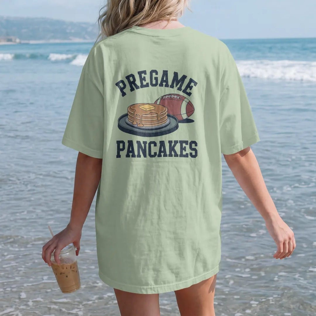 Woman on the beach in a Light Green tee, back view: a stack of pancakes on a camp griddle with butter and syrup, a football beside them, with "PREGAME" and "PANCAKES" in chunky 1970s swash script, printed in faded stadium green, washed brick red,…