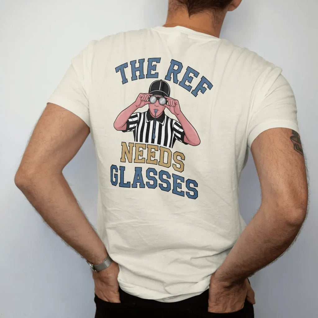 Man wearing a Ivory tee, seen from the back: a referee in stripes squinting through oversized glasses with a whistle in his mouth, with "THE REF" and "NEEDS GLASSES" in chunky rounded 70s varsity letters, printed in faded stadium green, washed…