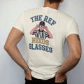 Man wearing a Ivory tee, seen from the back: a referee in stripes squinting through oversized glasses with a whistle in his mouth, with "THE REF" and "NEEDS GLASSES" in chunky rounded 70s varsity letters, printed in faded stadium green, washed…